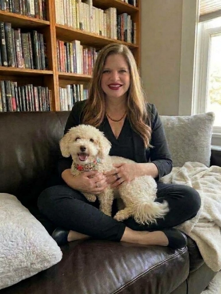 Dr. Karen McDowell sitting cross-legged on a leather couch in front of a bookshelf, holding Bennie, a fluffy cream-colored dog in a floral collar